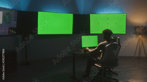 African American man works on computer in studio with equipment. Multiple chroma key big screen on the wall. Professional software displayed on tablet. Color grading or film editing room. Dolly shot.