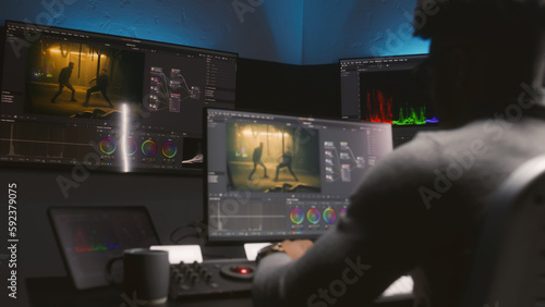 African American colorist makes color grading on control machine in modern studio. Big screens with action film footage and RGB colour correction graphic bar on the wall. Film post production process.