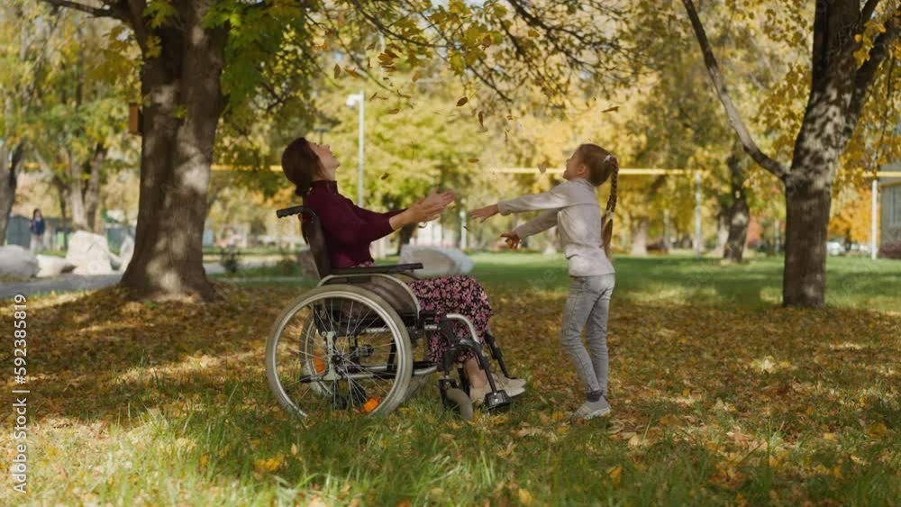 Mother with legs injury and daughter play in park. Girl throws up yellow leaves handful and couple waves hands looking upward joyfully. Loving family on vacation Mother with legs injury and daughter play in park. Girl throws up yellow leaves handful and couple waves hands looking upward joyfully. Loving family on vacation