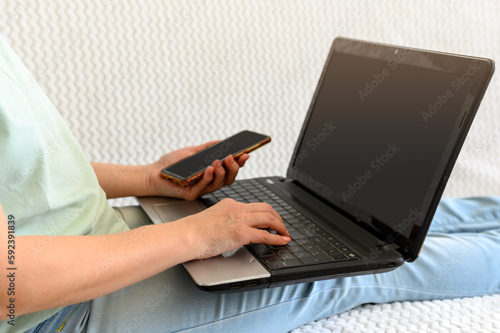 Fototapeta premium A girl with a phone in her hands is engaged at home on a laptop with a mug on the couch.