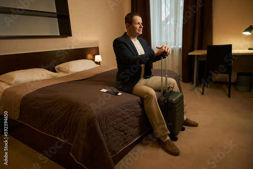 Male in travel clothes sits in bedroom of hotel room