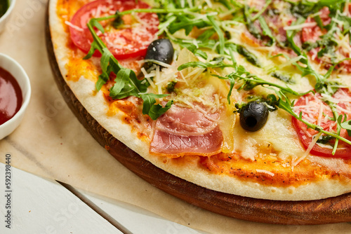 Freshly baked pizza with ham, rukkola, sauce pesto and olives served on wooden background with tomatoes, sauces and herbs. Food delivery concept. Restaurant menu