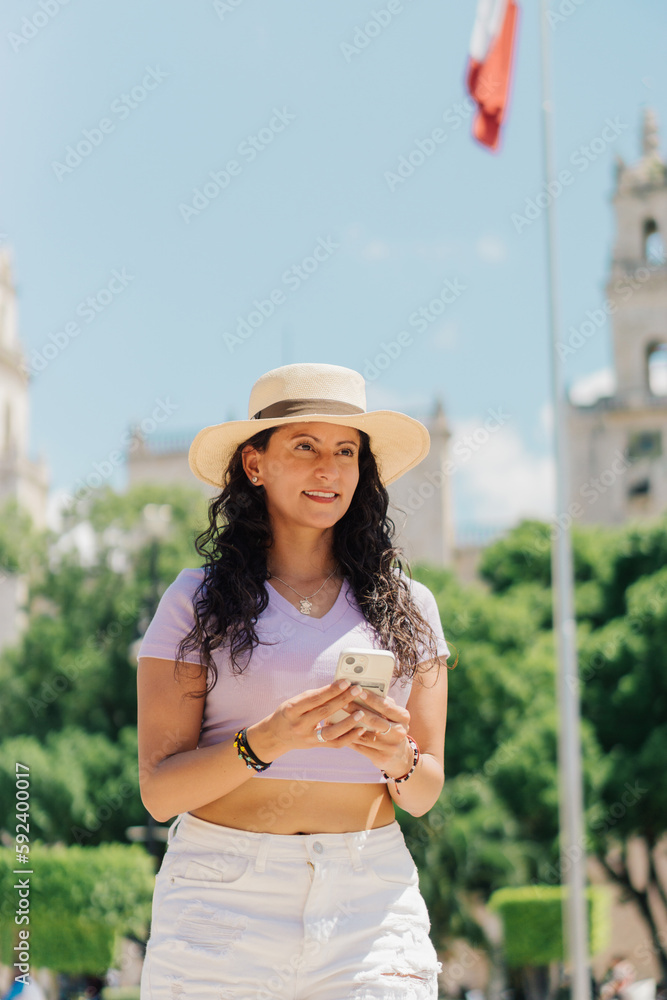 Latin woman holding a cell phone, walking and smiling on a sunny day.