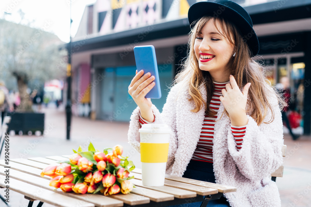 Stylish young smiling woman enjoying coffee from a reusable cup and having video call on her phone in a street cafe. Fresh tulips bouquet on the table. Springtime positive mood, street fashion.