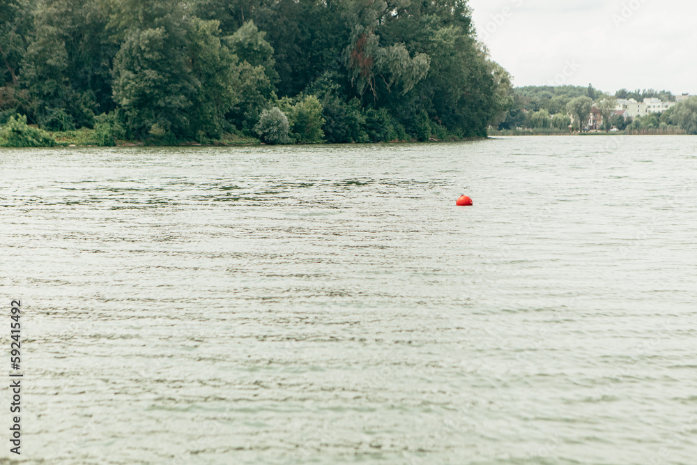 Red buoy in the water of lake sea or river in the city. Attention warning for human people, life saving, rescue floating device navigation. Summer day weather, island with green nature trees.
