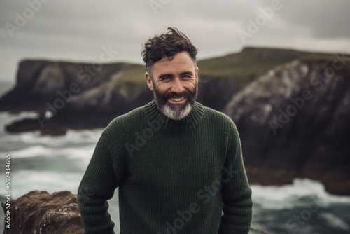 Portrait of a smiling bearded man in a green sweater standing on a cliff by the sea