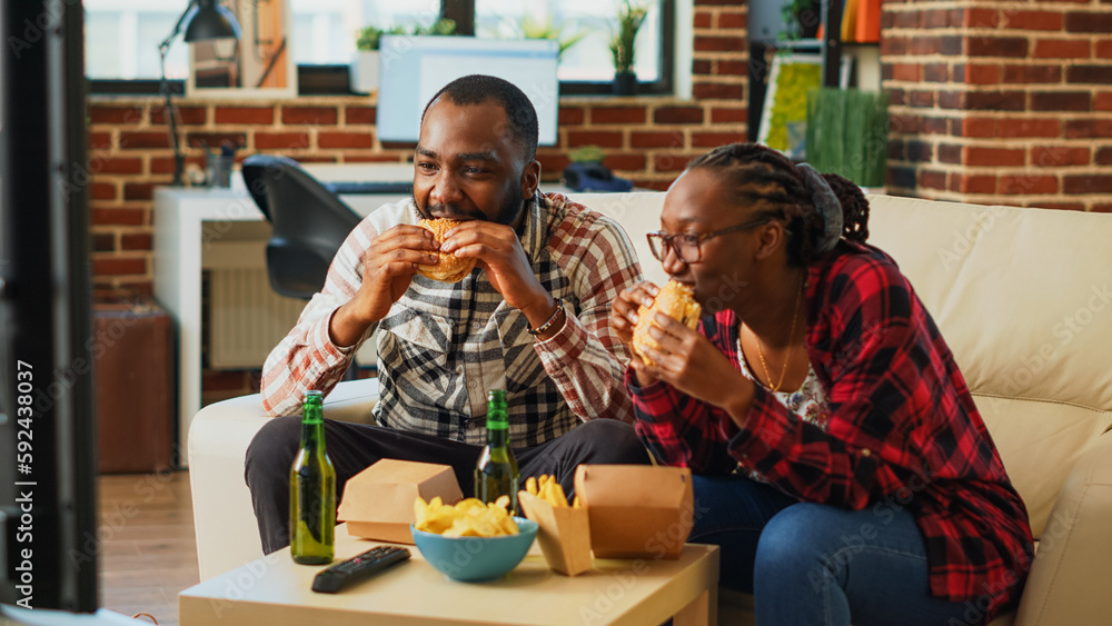 Obraz premium Modern young couple eating burgers together on couch, enjoying hamburgers with fries and bottles of alcohol. Cheerful man and woman having fun watching film on television, drinking beer.