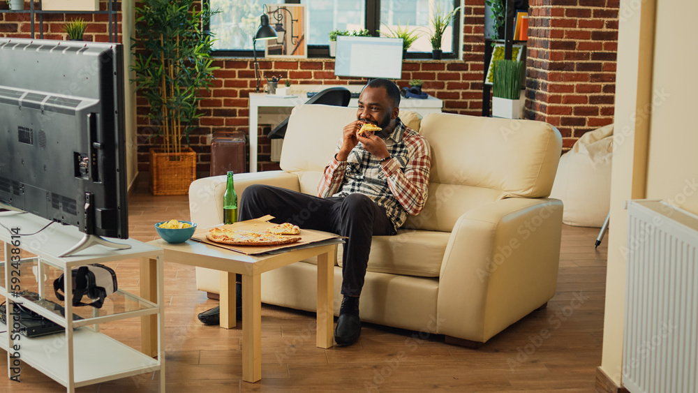 Cheerful adult taking bite of pizza at home, eating takeaway food delivery and drinking alcohol. Young relaxed man having fun watching action movie on television, eating snacks. Cheerful adult taking bite of pizza at home, eating takeaway food delivery and drinking alcohol. Young relaxed man having fun watching action movie on television, eating snacks.