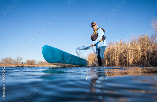 senior male paddler is rinsing his  stand up paddleboard after paddling on on a calm lake in early spring, frog perspective from an action camera