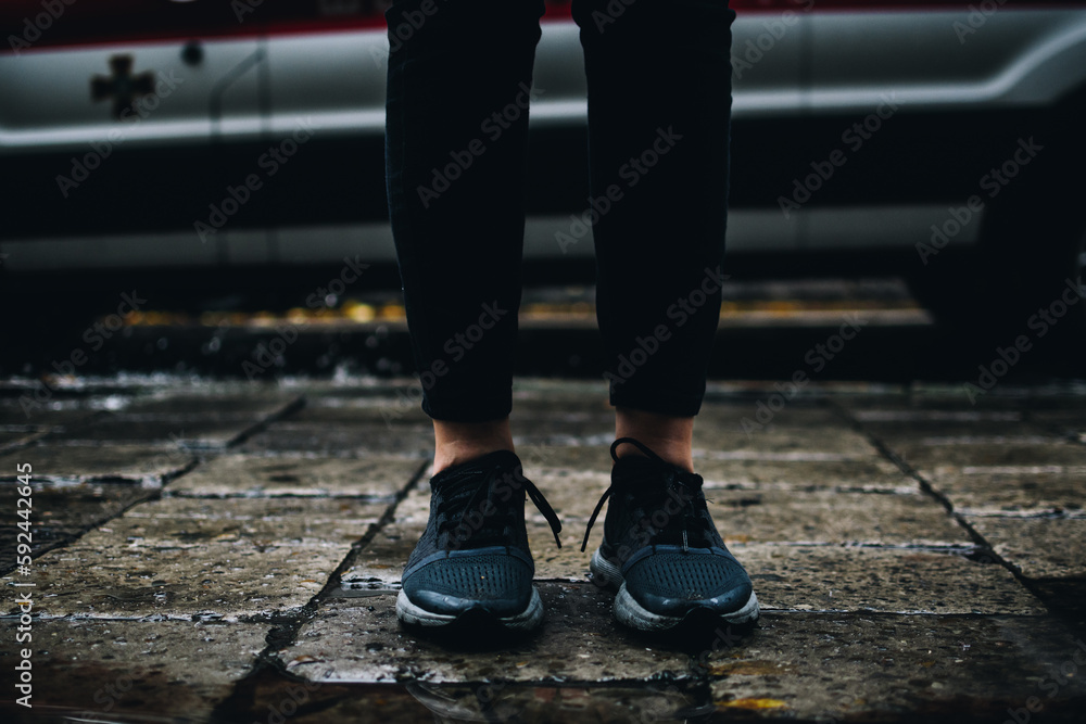 Girl wearing sneakers standing on the wet asphalt. Ambulance car and splashing puddle on the background.