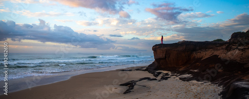 A beautiful girl in a pink tracksuit stands on a rock on Ten Mile Beach and enjoys a stunning orange sunrise over the Pacific ocean. Black rocks campsite, NSW, Australia