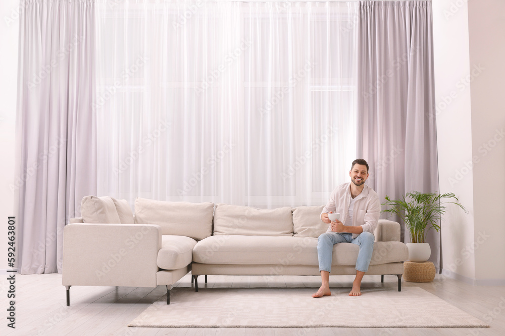 Happy man drinking coffee while resting on sofa near window with beautiful curtains in living room Happy man drinking coffee while resting on sofa near window with beautiful curtains in living room