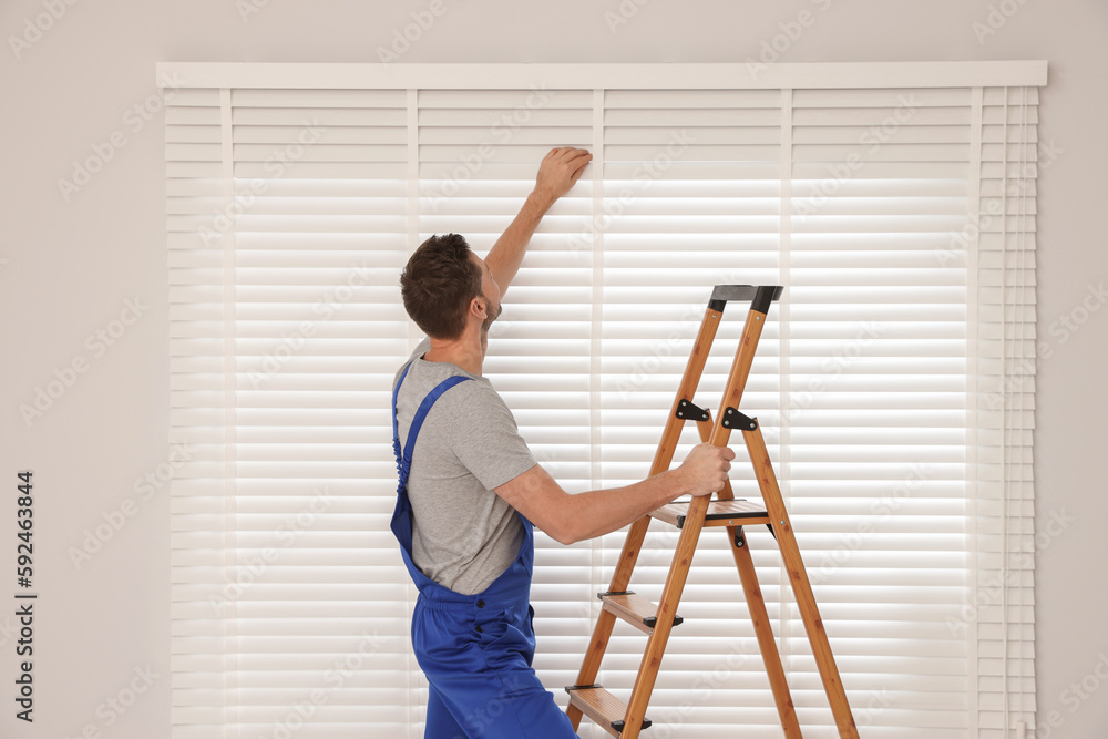 Worker in uniform installing horizontal window blinds on stepladder indoors Worker in uniform installing horizontal window blinds on stepladder indoors