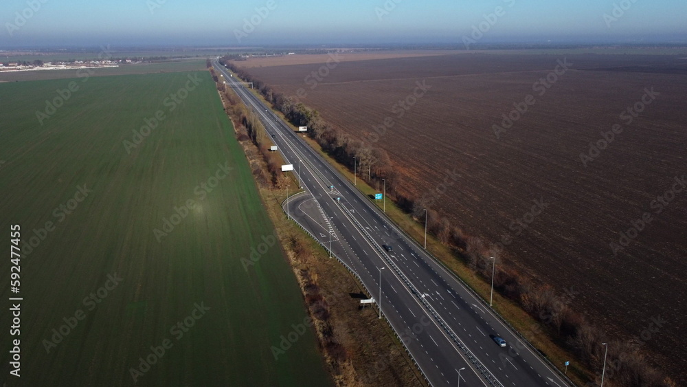Beautiful panoramic landscape. Black paved highway with white markings,many driving passing cars, large agricultural fields with green agricultural crops and plowed black soil on a sunny autumn day.