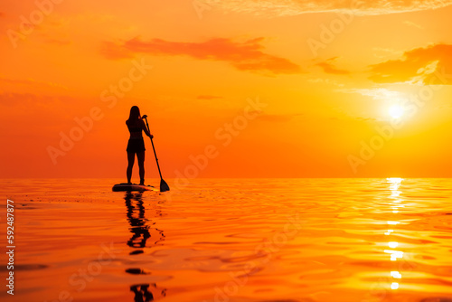 Silhouette of woman on stand up paddle board at sea with warm sunset or sunrise. Woman on SUP board and bright sunset with reflection on water