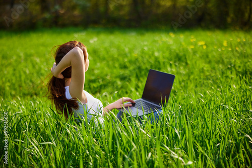a red-haired woman sits in a field of green grass with a laptop and straightens her long hair