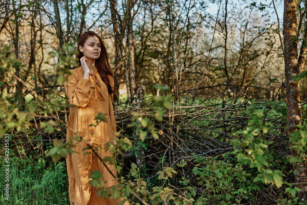 woman with long hair walks in the shade near the trees, dressed in a long orange dress, enjoying the weather and the weekend. The theme of privacy with nature, horizontal photography on the street woman with long hair walks in the shade near the trees, dressed in a long orange dress, enjoying the weather and the weekend. The theme of privacy with nature, horizontal photography on the street