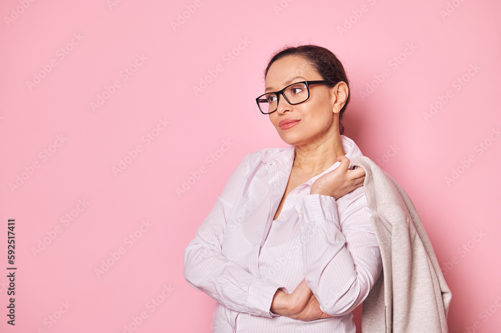 Dark-haired middle-aged pensive charming middle-aged business woman, wearing stylish glasses and casual clothes, holding her formal jacket and looking away over pink background. Copy advertising space Dark-haired middle-aged pensive charming middle-aged business woman, wearing stylish glasses and casual clothes, holding her formal jacket and looking away over pink background. Copy advertising space