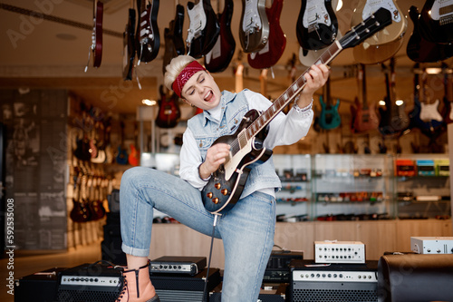 Woman rocker playing guitar connected to amplifier over shop store background