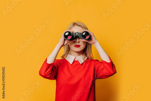 Stylish blonde hair woman in red dress with binoculars on yellow background