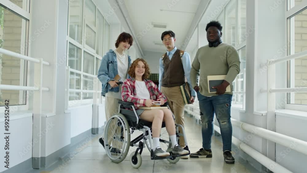 Long shot of group of modern multi-ethnic students with girl with disability posing on camera in college corridor Long shot of group of modern multi-ethnic students with girl with disability posing on camera in college corridor