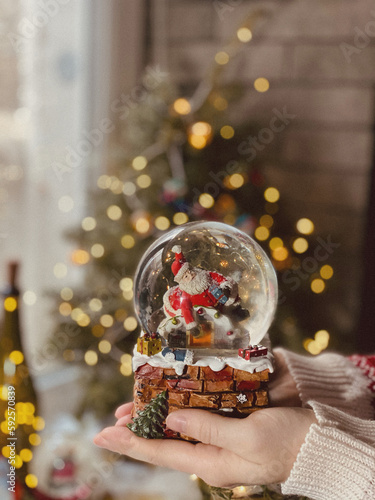 Magic snow globe with a figurine of Santa Claus with deers and Christmas decorations on the background of the Christmas tree.Bokeh lights. snow globe in female hands.concept of new year and christmas.