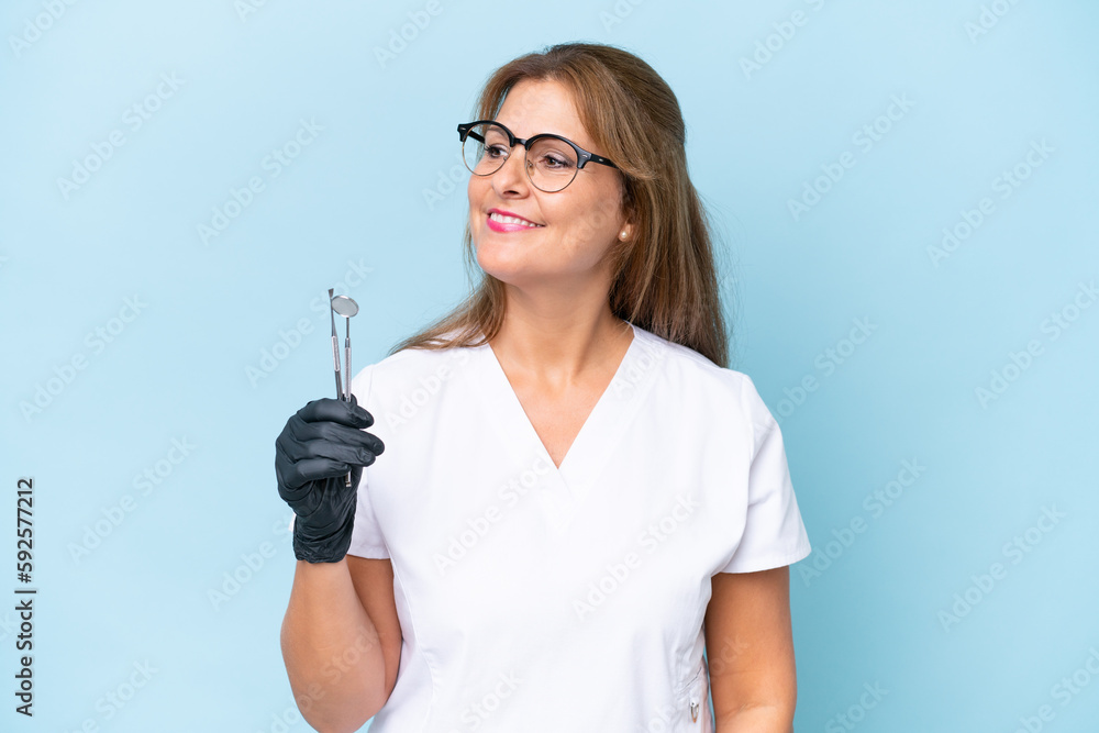 Middle-aged Dentist holding tools over isolated blue background looking to the side and smiling Middle-aged Dentist holding tools over isolated blue background looking to the side and smiling