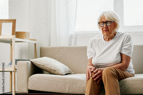 Elderly woman severe pain in her leg sitting on the couch, health problems in old age, poor quality of life. Grandmother with gray hair holds on to her sore knee, problems with joints and ligaments. © SHOTPRIME STUDIO