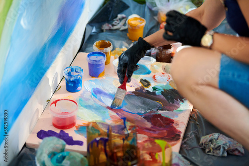 Artist hand in black gloves holds paint brush and choose color from colorful palette at outdoor art painting festival, paintings art picture process. Artist paints atmospheric surreal picture photo