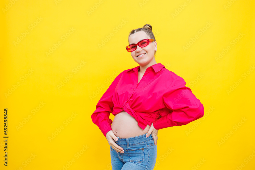 Happy pregnant woman with a smile on her face. Cheerful pretty pregnant woman in a pink shirt and pink glasses on a yellow background. Young bright pregnant woman.