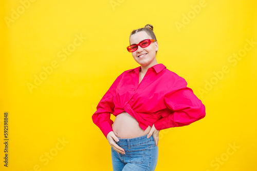 Happy pregnant woman with a smile on her face. Cheerful pretty pregnant woman in a pink shirt and pink glasses on a yellow background. Young bright pregnant woman.