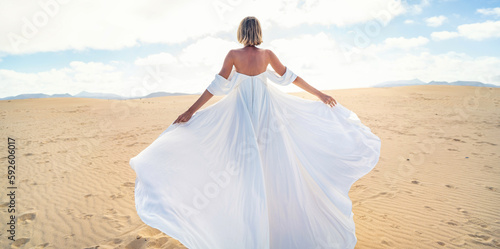 Back view of a blonde woman in fashionable, maxi white wedding dress posing on the desert, dancing in sunny light.