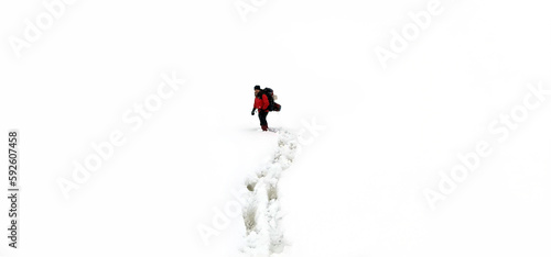 A man in a red jacket with a backpack walking on snow, footprints in snow, behind.
