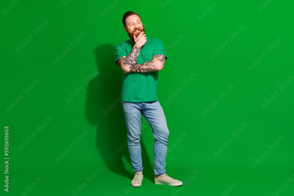 Full length photo of cheerful dreamy guy dressed t-shirt smiling arm chin looking empty space isolated green color background Full length photo of cheerful dreamy guy dressed t-shirt smiling arm chin looking empty space isolated green color background