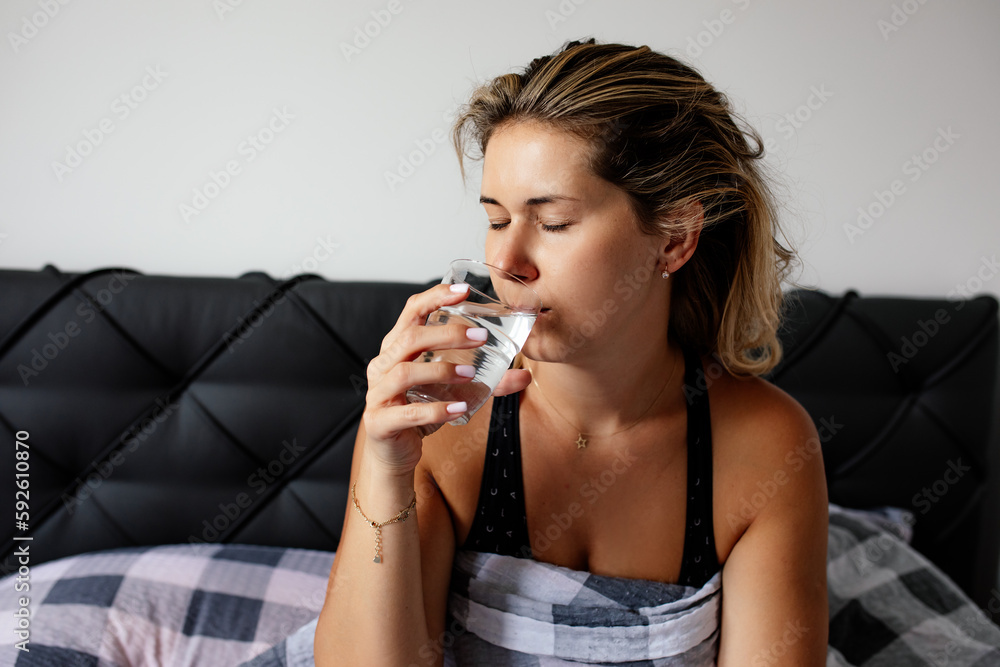 Young woman still not fully awake, with her eyes closed, drinks water sitting in bed in morning. Proper nutrition. Young woman still not fully awake, with her eyes closed, drinks water sitting in bed in morning. Proper nutrition.