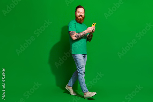 Full length photo of positive funky man wear t-shirt texting messages modern gadget isolated green color background