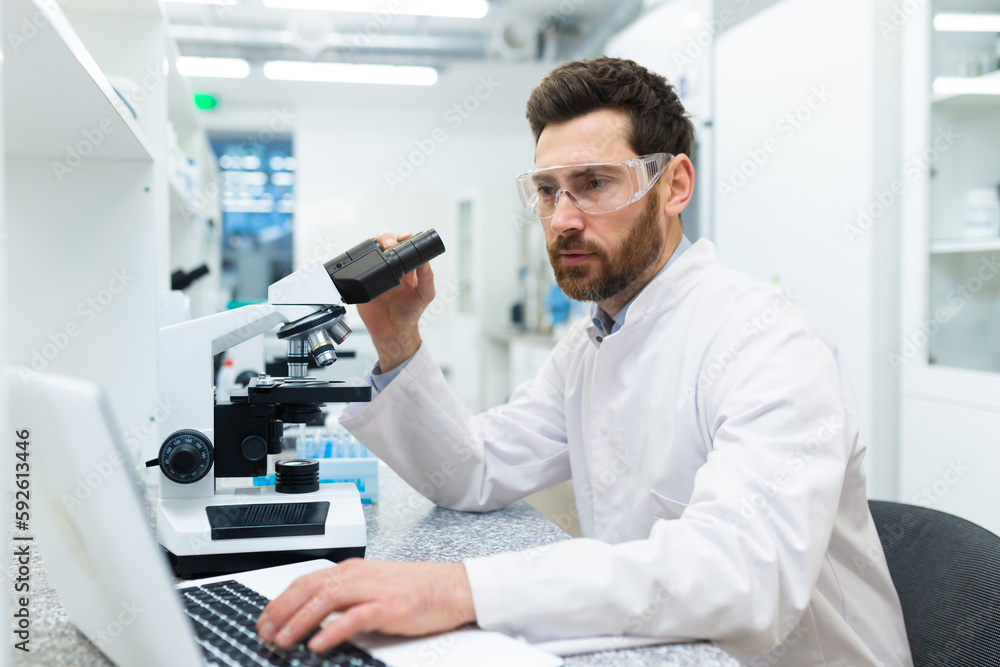 Serious and thoughtful scientist working in laboratory with microscope and laptop, mature experienced man in glasses and medical gown conducting experiments and entering data online into computer .