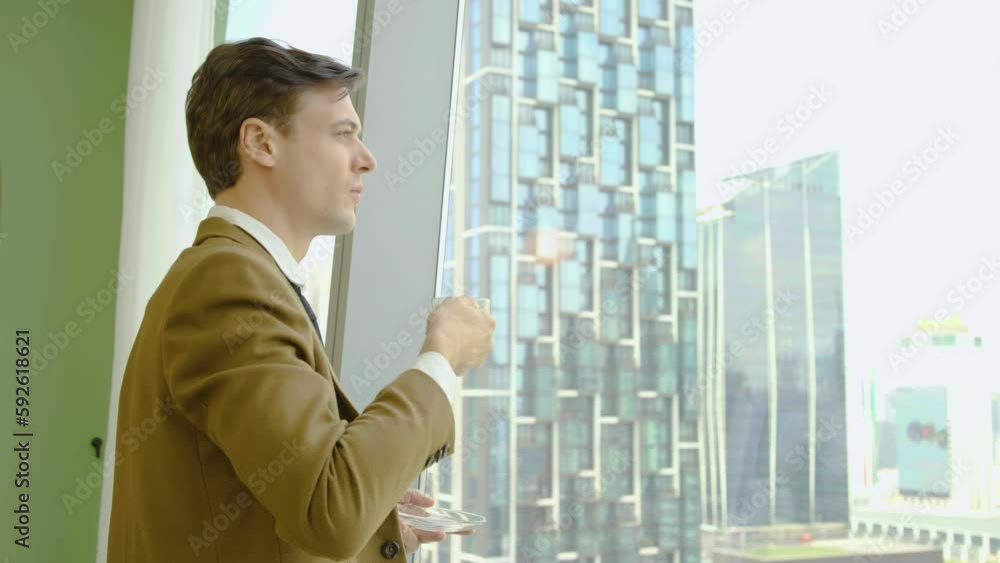 Young caucasian businessman drinking coffee or tea and admire view standing by window in office, enjoying beautiful morning feeling happy after successful deal signing contract profitful partnership Young caucasian businessman drinking coffee or tea and admire view standing by window in office, enjoying beautiful morning feeling happy after successful deal signing contract profitful partnership