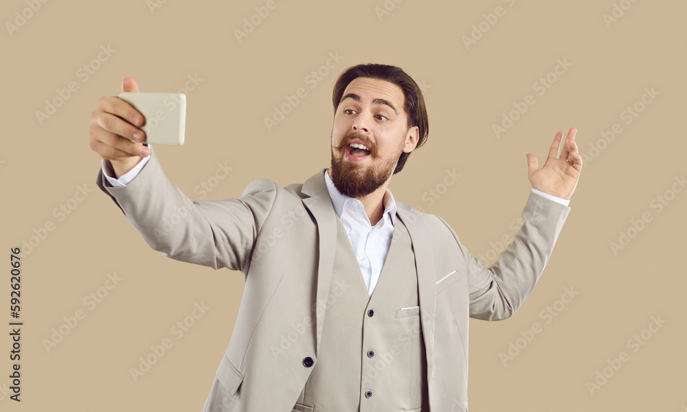 People lifestyle. Cheerful stylish man in suit takes selfie, records video or talks online on beige background. Smiling active millennial Caucasian man in three-piece suit uses modern smartphone. People lifestyle. Cheerful stylish man in suit takes selfie, records video or talks online on beige background. Smiling active millennial Caucasian man in three-piece suit uses modern smartphone.