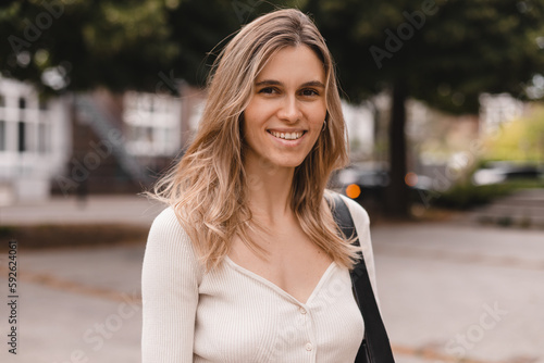 Close up portrait attractive young happy woman with fresh and clean skin stands outside city in park. Smiling curly blonde wear long sleeves top and look at camera. Lifestyle, female beauty concept.