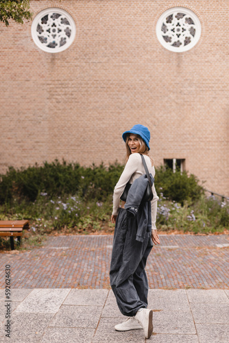 Full length image of radiant young caucasian girl walking against street wall. Blonde woman is wearing wide pants cargo, sweater top with buttons and blue panama hat, bag on shoulder, she turn around.