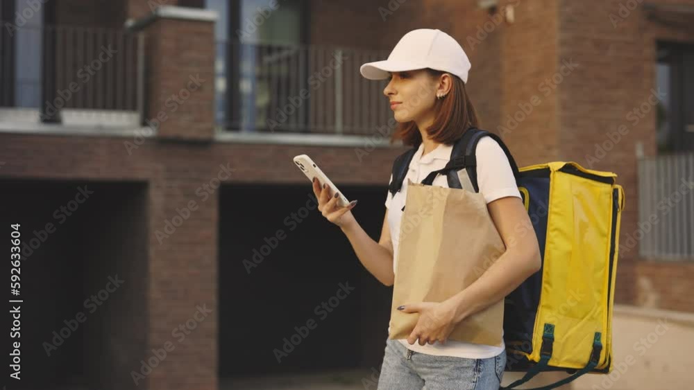 Courier Going Along Building Holding the Paper Package and Mobile Phone in Hand. Delivery Lady Looking for Delivery Address on Street. Cute Woman Using Smartphone While Food Delivering Outdoors Courier Going Along Building Holding the Paper Package and Mobile Phone in Hand. Delivery Lady Looking for Delivery Address on Street. Cute Woman Using Smartphone While Food Delivering Outdoors