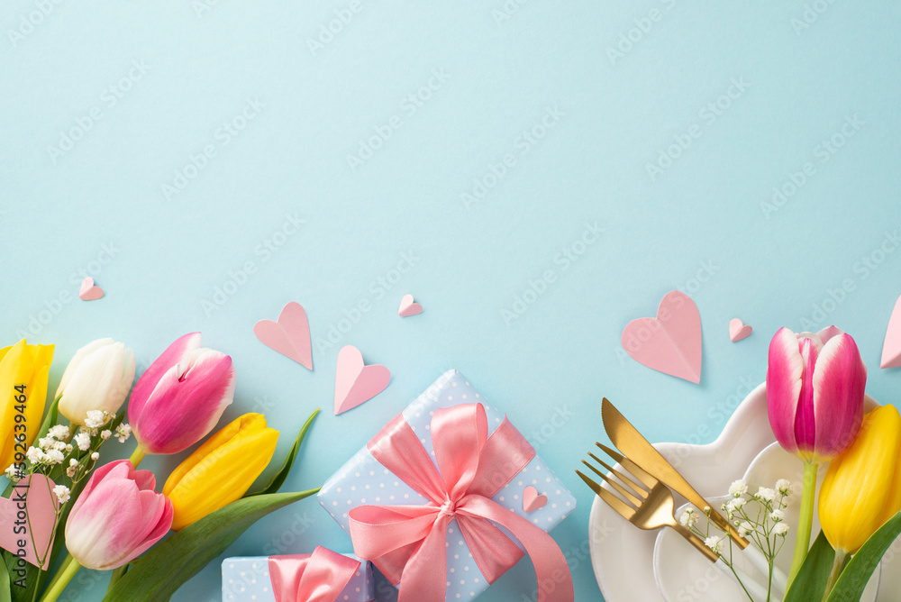 Mother's day concept. Top view flat lay of trendy table setting featuring plates, cutlery, tulips, gift boxes and decorative hearts on pastel blue background. Empty space for text or advert