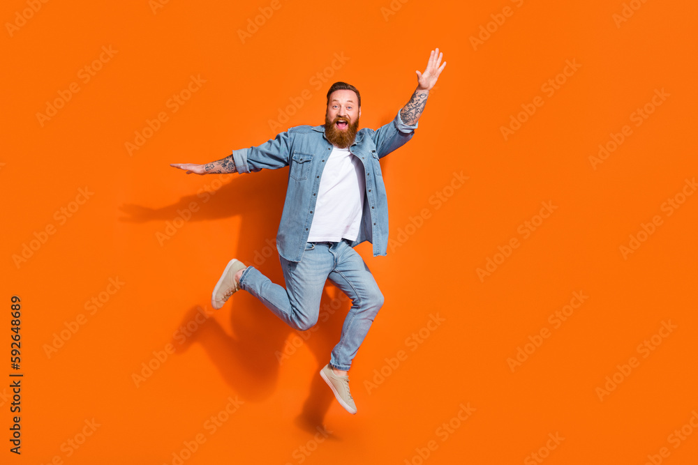 Full length photo of cheerful funky guy dressed denim jacket jumping high empty space isolated orange color background
