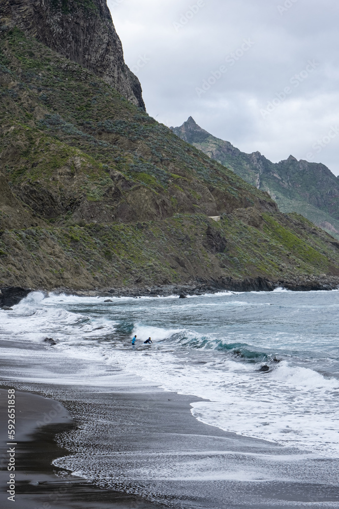 People surfing alone in a black sand beach with waves surrounded by nature and mountains in cloudy day in tenerife, canarias, spain People surfing alone in a black sand beach with waves surrounded by nature and mountains in cloudy day in tenerife, canarias, spain