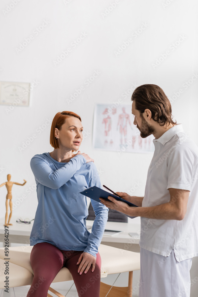 Fototapeta premium woman touching painful shoulder during consultation with physiotherapist holding clipboard in consulting room.