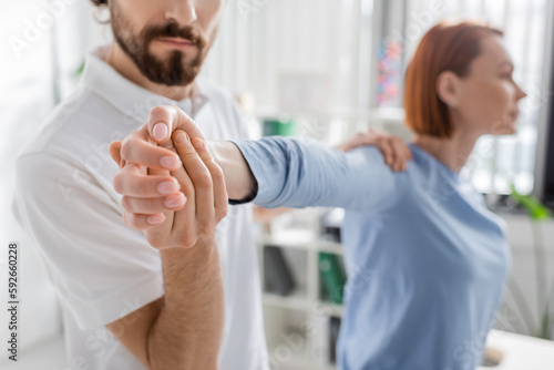 bearded manual therapist examining injured arm of blurred woman in consulting room.