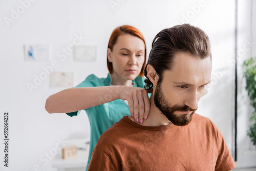 blurred osteopath touching painful neck of bearded man during examination in consulting room.