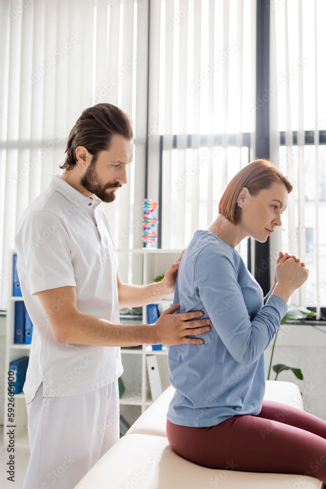 side view of bearded physiotherapist touching painful back of redhead woman during appointment in consulting room. side view of bearded physiotherapist touching painful back of redhead woman during appointment in consulting room.