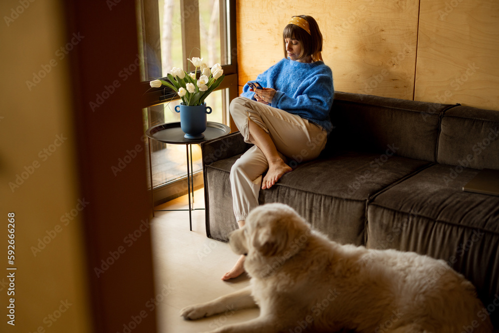 Woman sitting relaxed with a phone on a couch by the window at wooden cottage. Concept of home comfort and leisure time Woman sitting relaxed with a phone on a couch by the window at wooden cottage. Concept of home comfort and leisure time